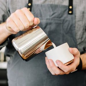 Barista gießt Milch aus einer polierten Kanne aus Edelstahl, Teil von Espresso Zubehör und Milchkännchen, alles rund um Kaffee.