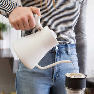 Hand, die Wasser aus einem weißen Wasserkocher mit schmalem Ausguss in einen Kaffeefilter gießt, Teil von EspressoGear, alles rund um Kaffee.